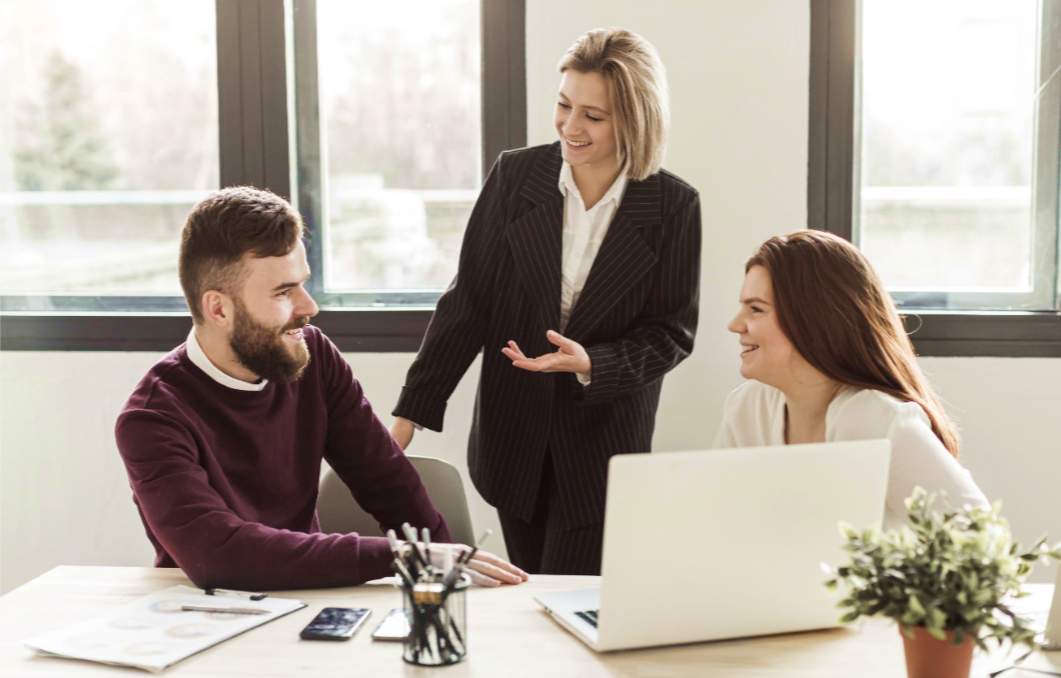 Trois personnes en tenue professionnelle échangent autour d’une table de bureau, avec un ordinateur portable, des documents, un pot à crayons et une plante verte, dans une pièce lumineuse avec de grandes fenêtres.